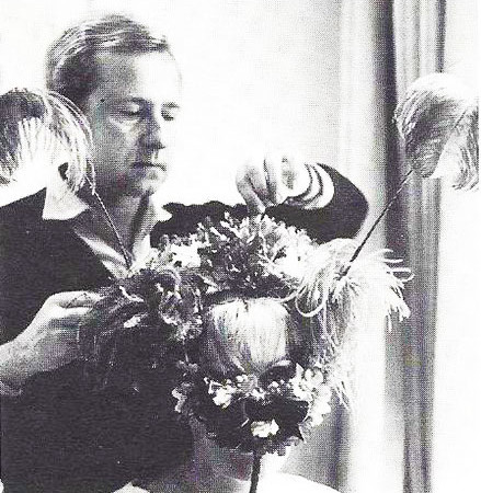 Originally a Milliner, Adolfo was formed his fantasy creations. He is shown adding the finishing touches to a 1965 confection of violets and feathers.